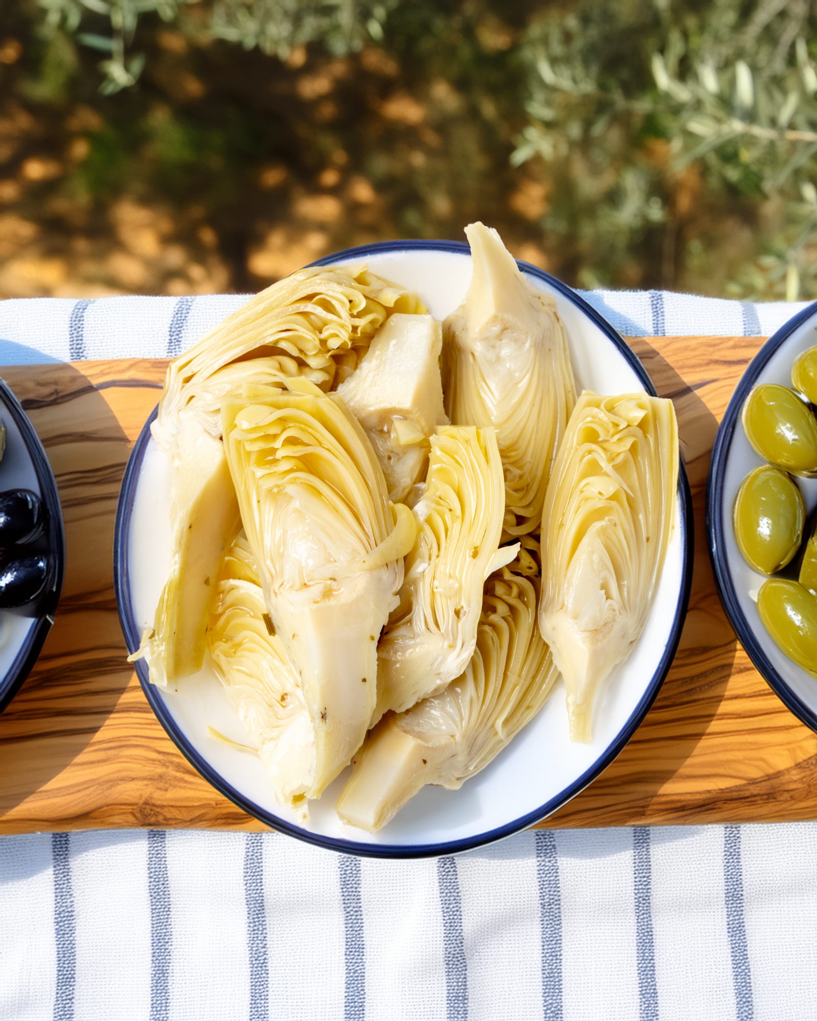 Italienische Artischockenherzen in Kräuteröl im Glas – serviert auf einem Teller, im mediterranen Gartenambiente mit Oliven und frischem Gemüse. Perfekt als Antipasti oder Beilage.